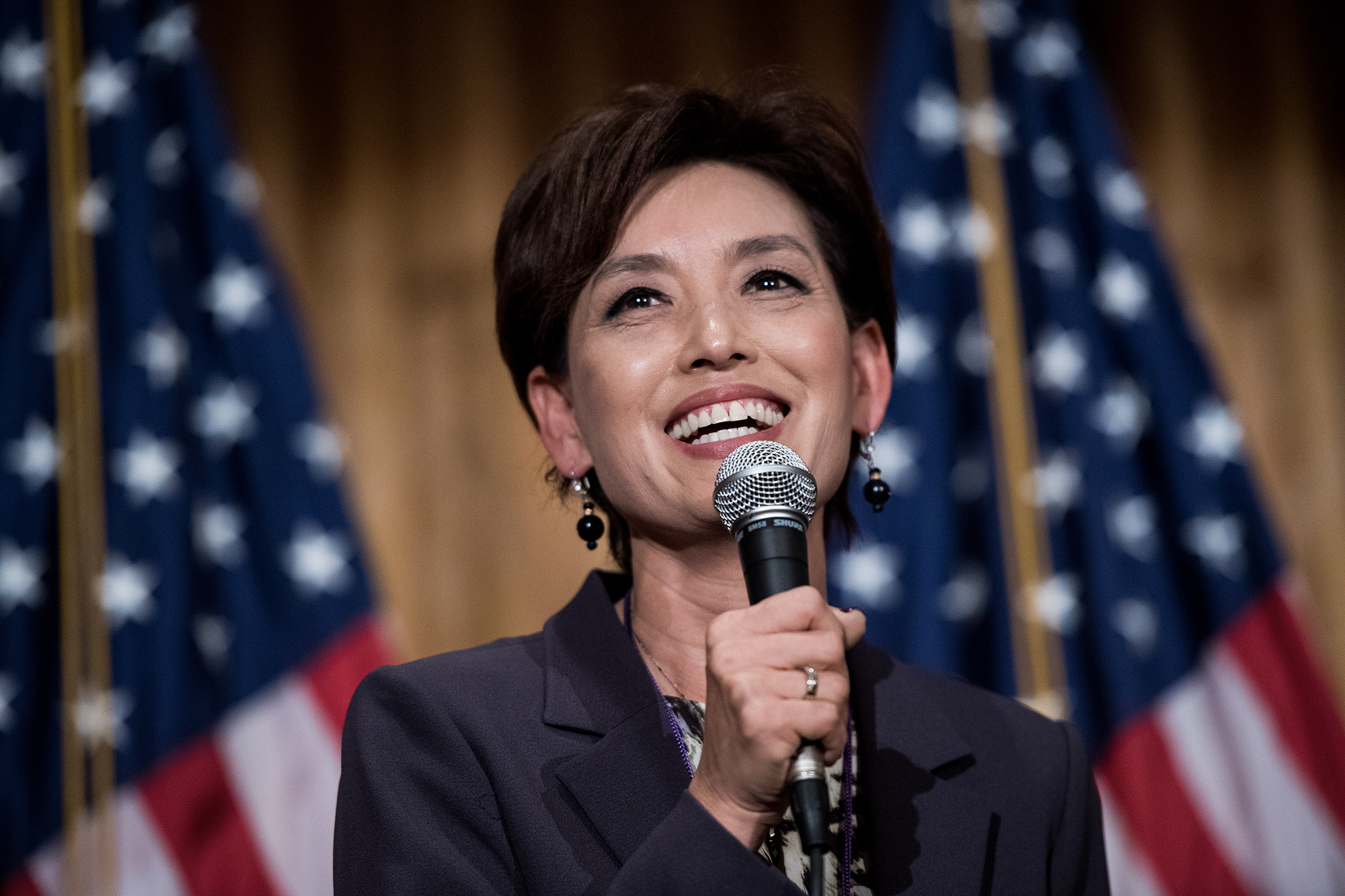 Most California Republicans in Congress won’t commit to certifying the 2024 presidential election In this close image, a smiling person, dressed in a suit, holds a microphone towards their face. Behind them, and out of focus, two American flags are visible. The scene is a forum and a discussion is taking place.