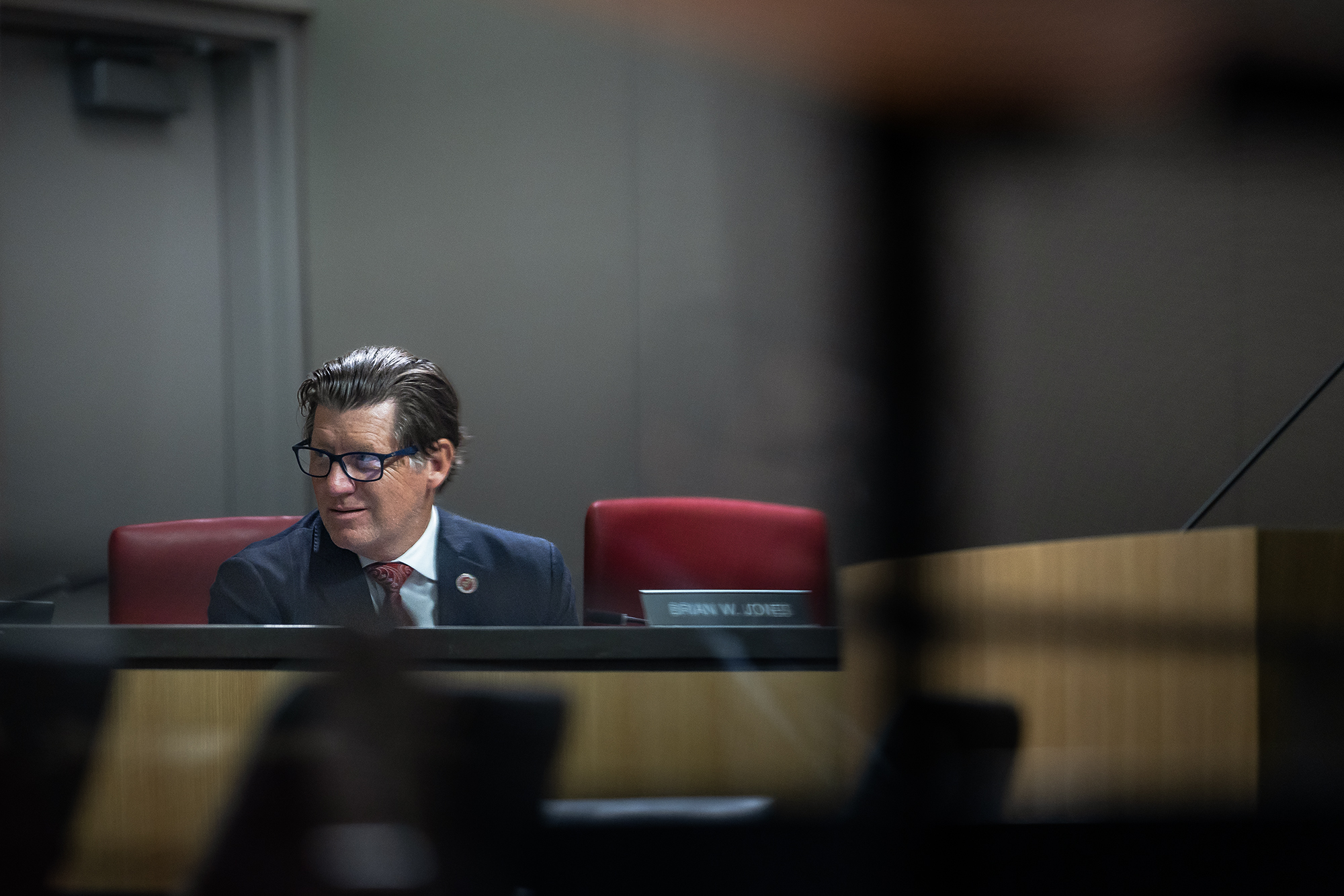 He wants to make California Republicans relevant again. So why does the GOP dislike him? A person seated in a formal meeting room is captured through a blurred foreground. The individual wears glasses, a suit, and a tie, seated on a red chair with a nameplate visible on the desk. The neutral-toned walls and wooden furniture add to the professional atmosphere.