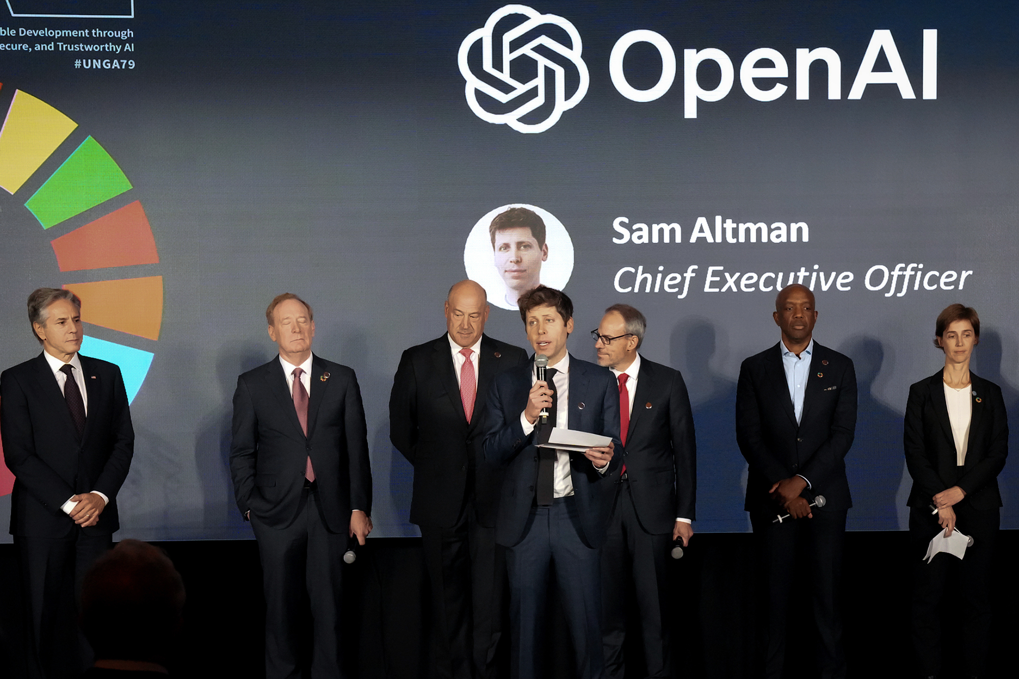 California is investigating OpenAI’s conversion to a for-profit company A group of individuals standing on a stage, dressed in formal attire, with one person holding a microphone and speaking. The background displays the "OpenAI" logo, colorful graphic elements, and text identifying "Sam Altman, Chief Executive Officer." The setting suggests a formal event or presentation.