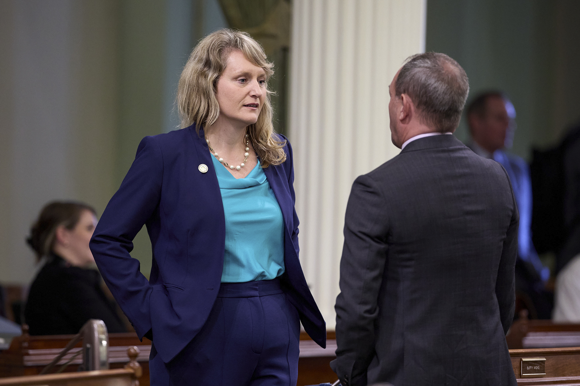 How Newsom has responded to the LA fires A person wearing a blue suit stands up and speaks with another person facing them in a grey suit while they stand in the Capitol assembly floor.