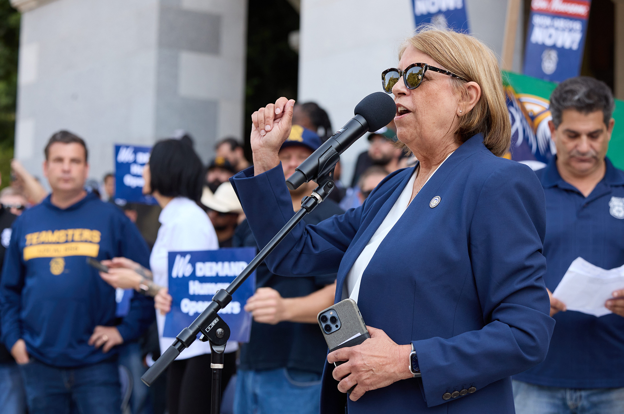Zombie bills: Why California lawmakers bring back legislation governors kill A person in a blue suit and sunglasses speaks into a microphone in front of a crowd of people standing on a set of stairs and holding signs outside the state capitol building.