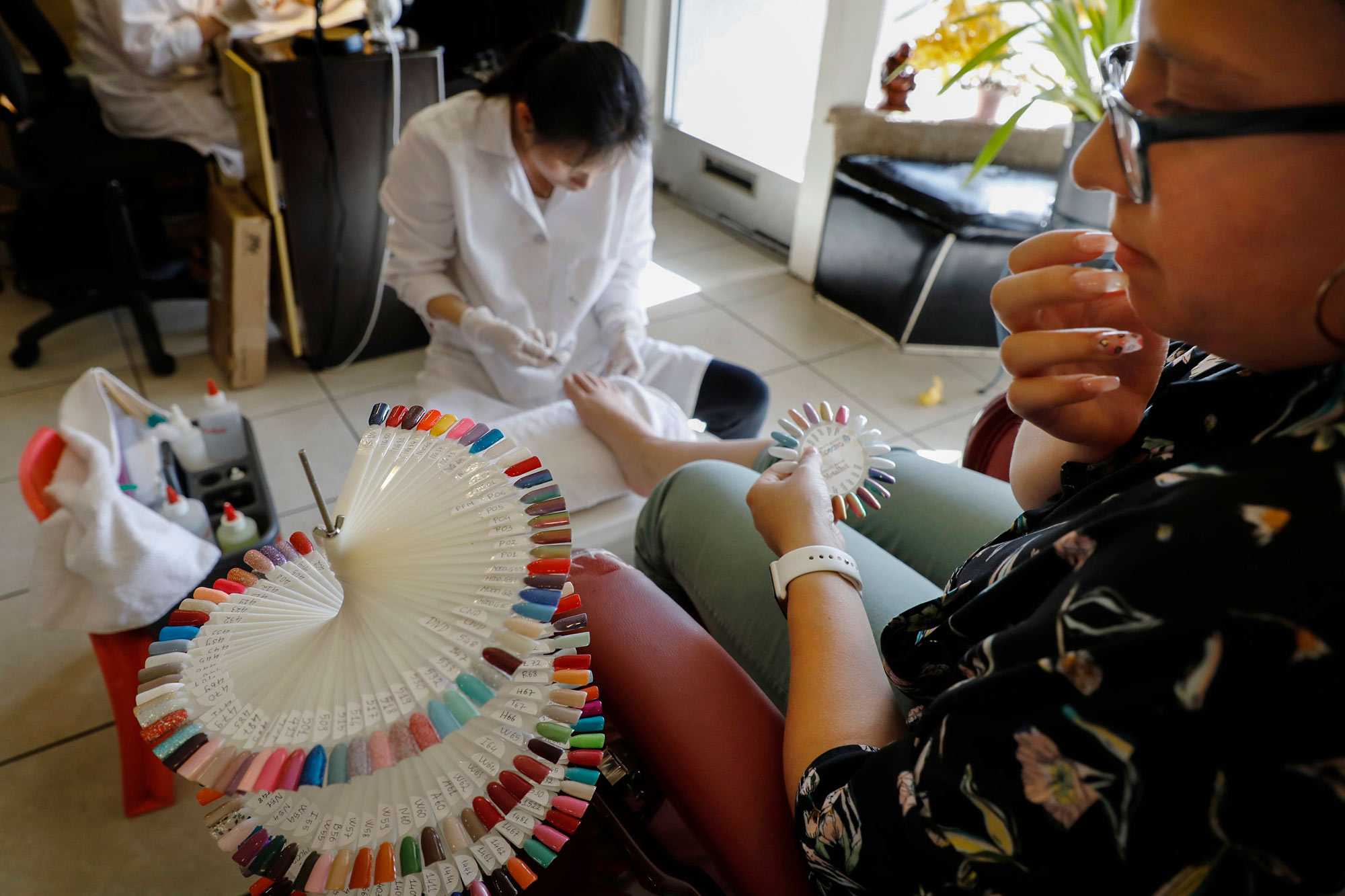 Why nail salons are still being targeted in fights over California gig worker law A person places their right hand on their chin while they contemplate which nail color they should get based on a nail display in front of them. A nail technician can be seen in the background giving the same person a pedicure.