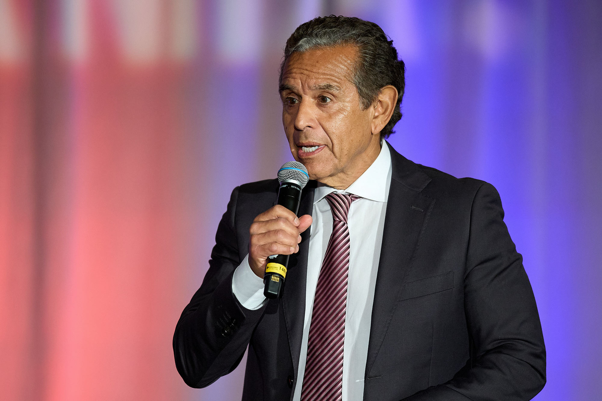 Newsom gave his political rival a $380k job. See the special interests who paid for it A person — with short, curly, dark hair and wearing a black suit with a pink pattern tie — holds a microphone while speaking at an event. The background is illuminated with soft pink and purple lights.