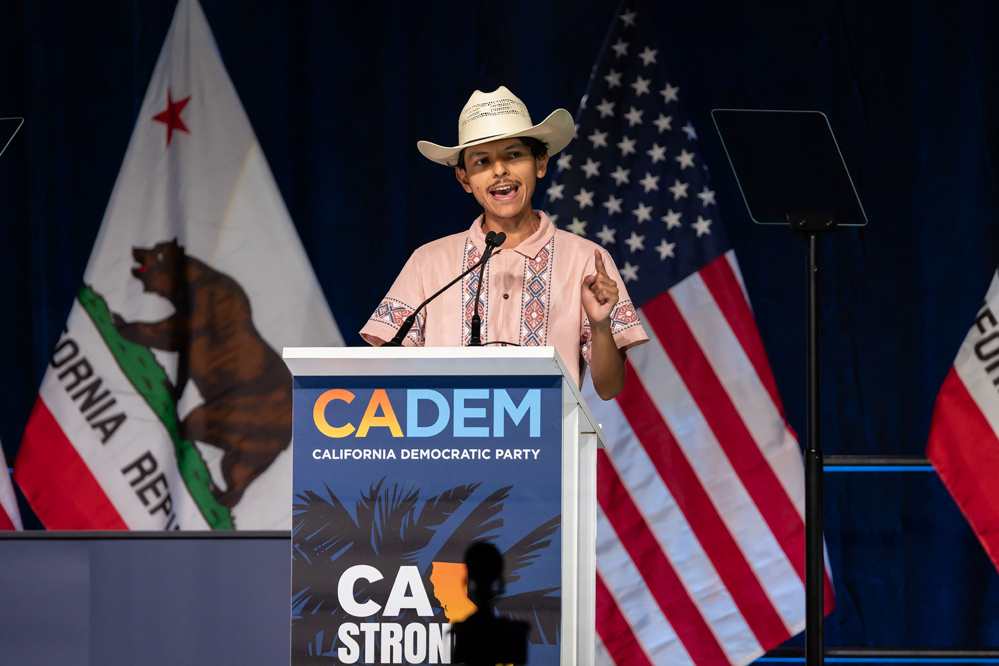 Young Democrats offer lessons for their leaders at party convention A person wearing a cowboy hat stands in front of a lectern with a sign in front of it that reads "California Democratic Party." The California state and American flags can be seen behind the person.
