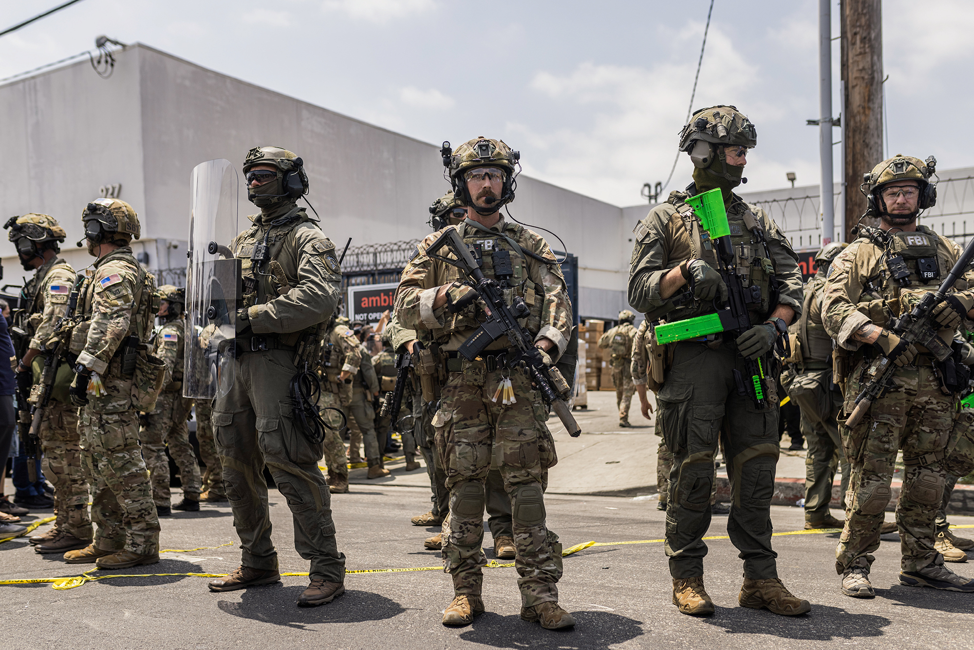 Worksite immigration raids are supposed to free up jobs for citizens. Here’s what really happens A line of heavily armed federal officers in tactical gear stands on a city street during a protest. Some wear FBI patches and hold rifles, one of which has a neon green magazine. Yellow police tape marks the ground, and a barbed-wire fence and industrial building labeled "ambiance" are visible in the background. The officers face forward, maintaining a defensive posture.
