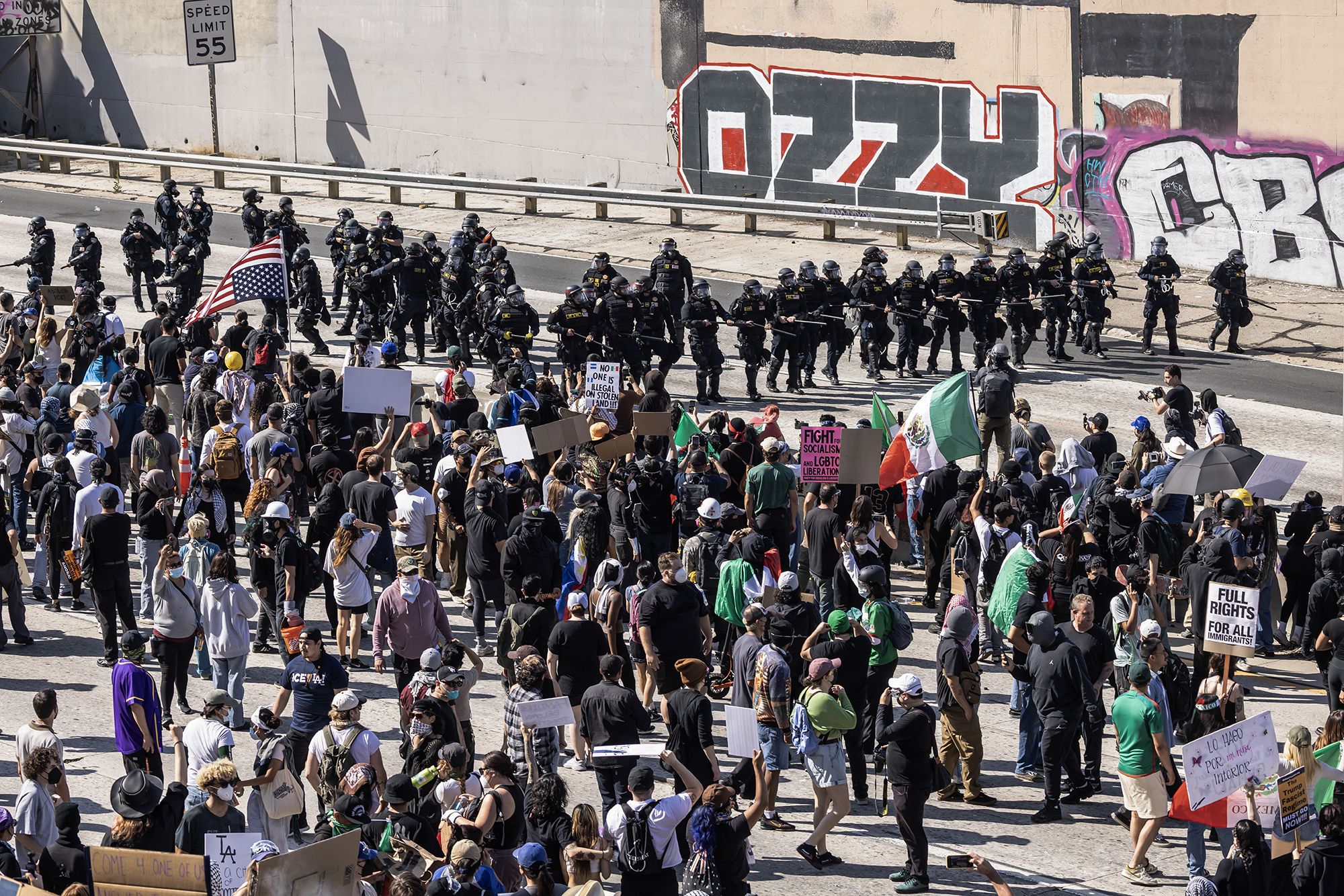 Newsom-Trump truce explodes as president deploys troops against his wishes A large crowd of protesters holding signs and flags, including Mexican and American flags, gathers on a street during a demonstration. Opposite them, a line of law enforcement officers in full riot gear stands shoulder-to-shoulder, forming a barrier. Graffiti covers the walls behind the officers, and the scene is marked by tension and confrontation.