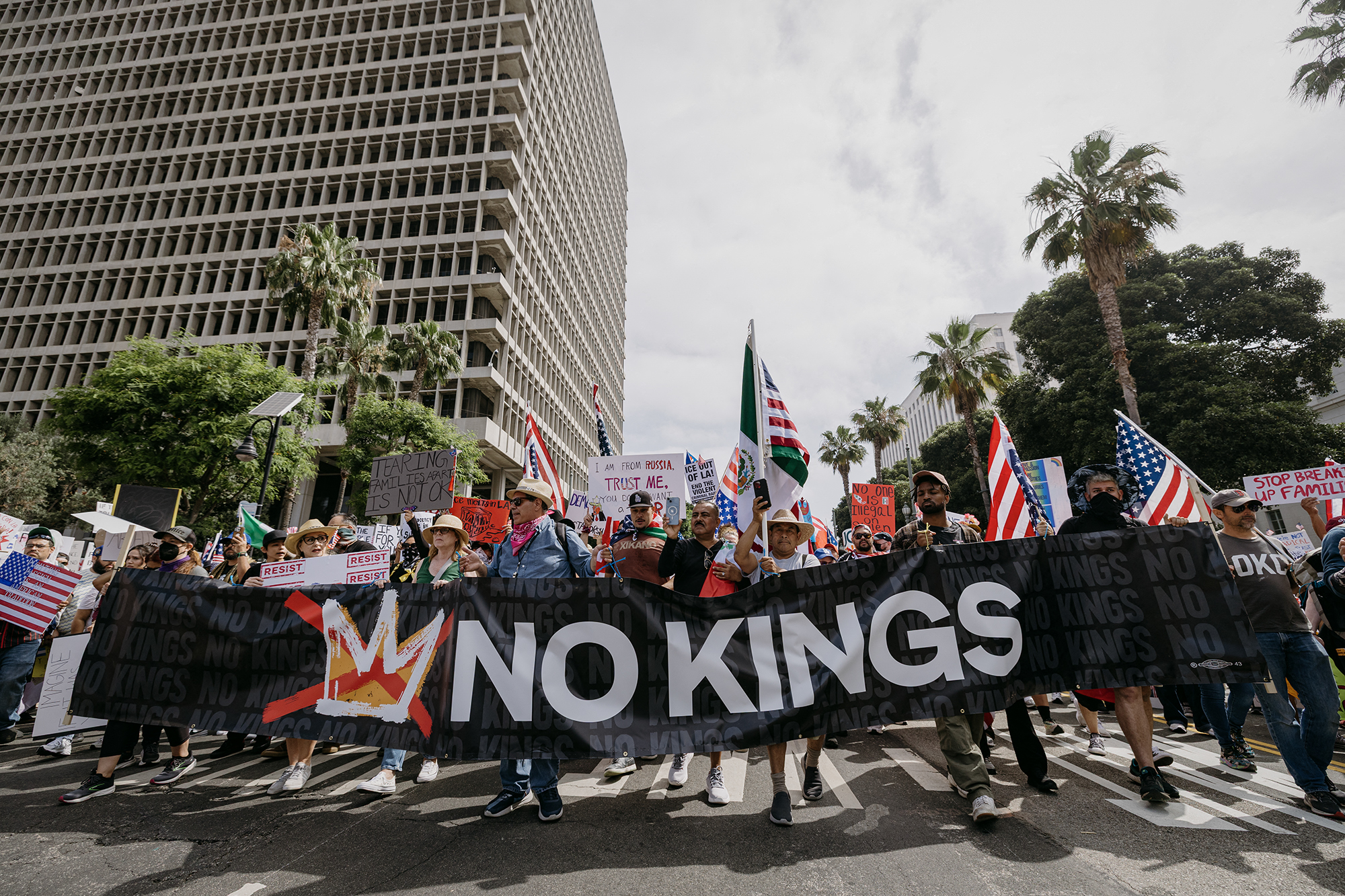 Trump lawyers call his decision to send troops to LA ‘unreviewable’ by U.S. courts A large group of demonstrators march down a city street holding a black banner that reads "NO KINGS" in bold white letters. Protesters carry American flags, handwritten signs, and wear hats and bandanas. Tall office buildings and palm trees frame the background under a cloudy sky.