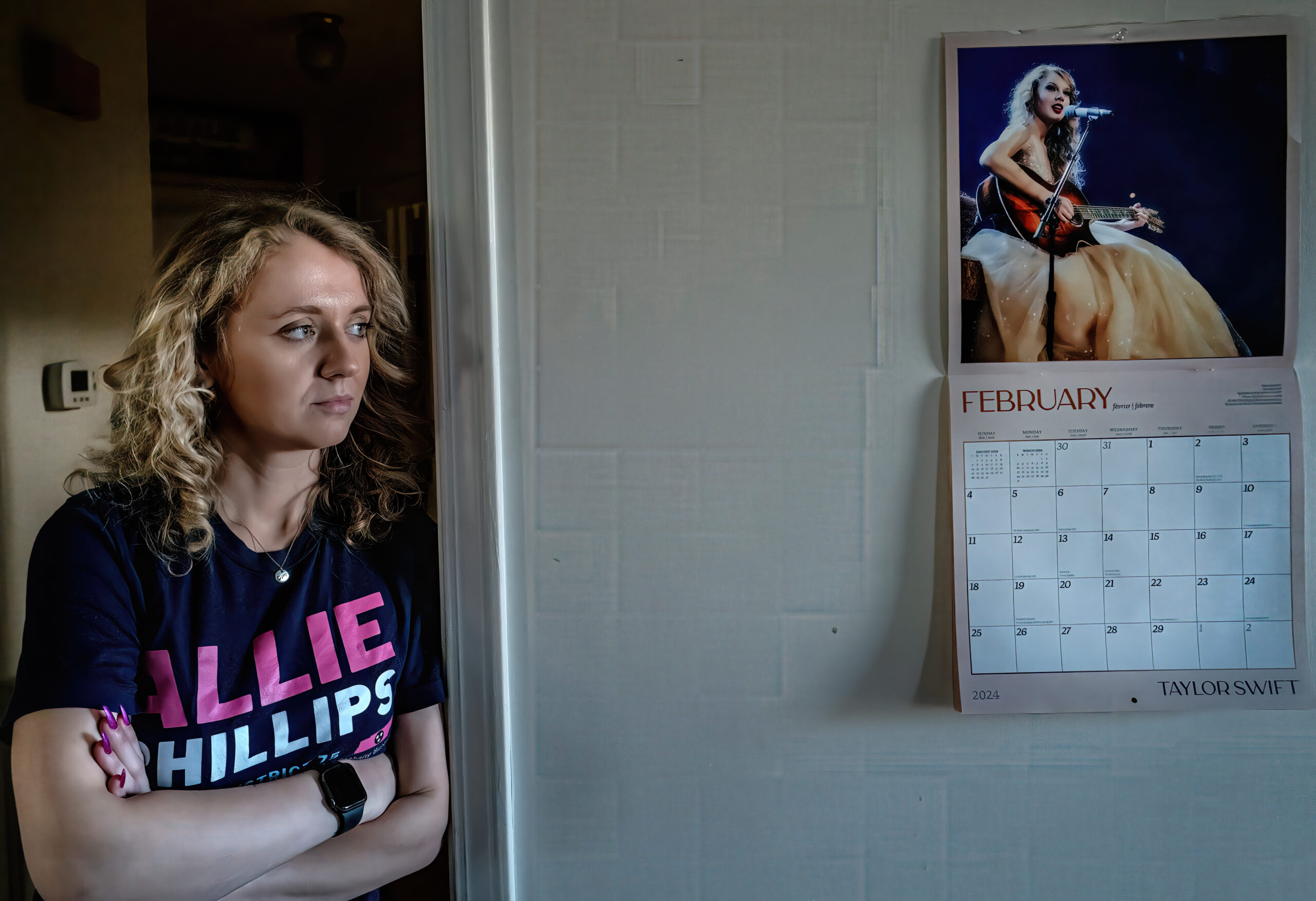 Turning pain into purpose: Tennessee House candidate talks to voters about her abortion A woman in a shirt saying "Allie Phillips" leaning against a wall with a calendar of Taylor Sewift visible