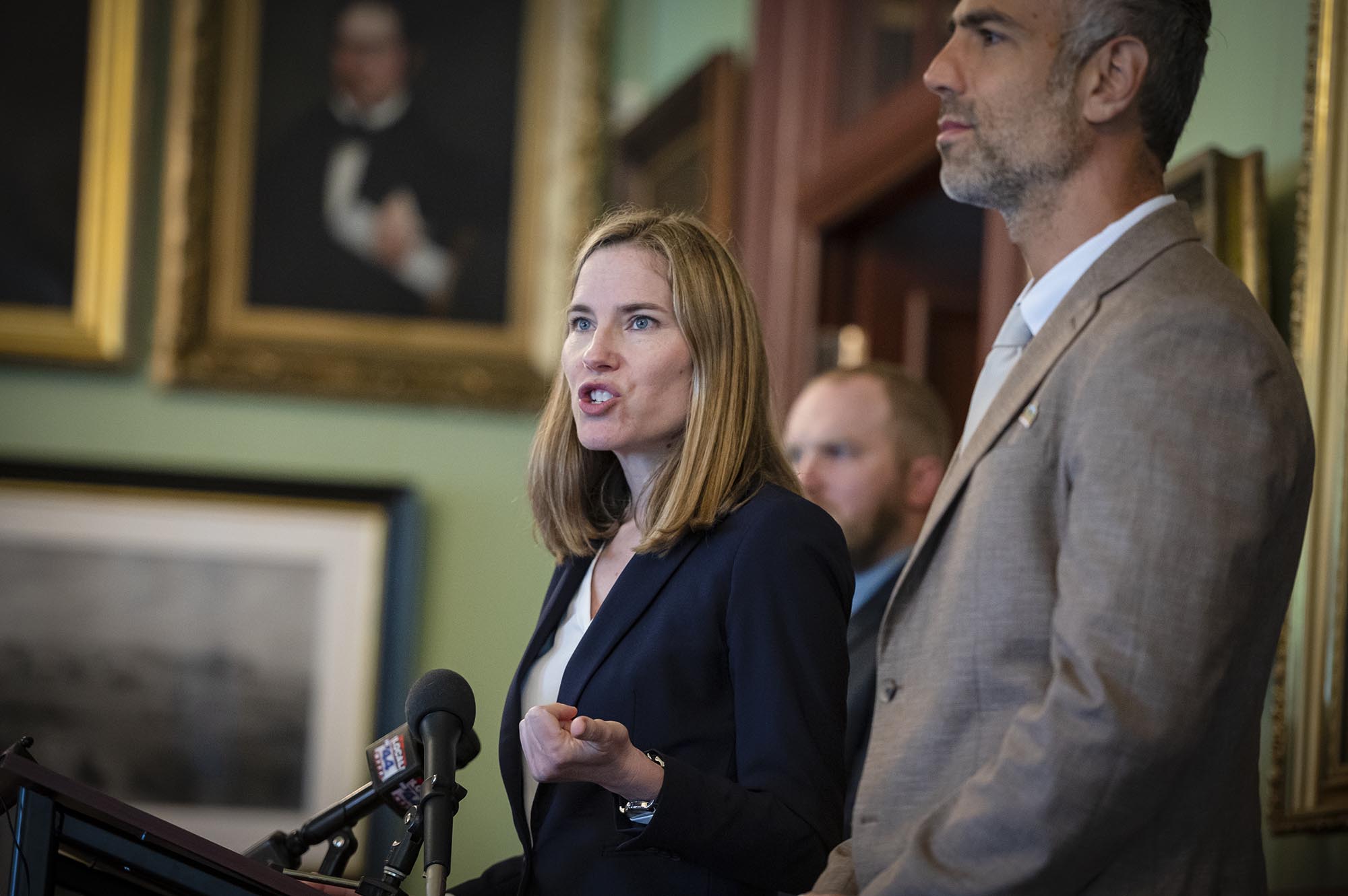 Amazon to pay $400,000 in settlement over Vermont ban on tobacco product sales A woman in a dark blazer speaks at a podium with microphones, while a man in a light suit stands beside her. Portraits hang on the wall in the background.