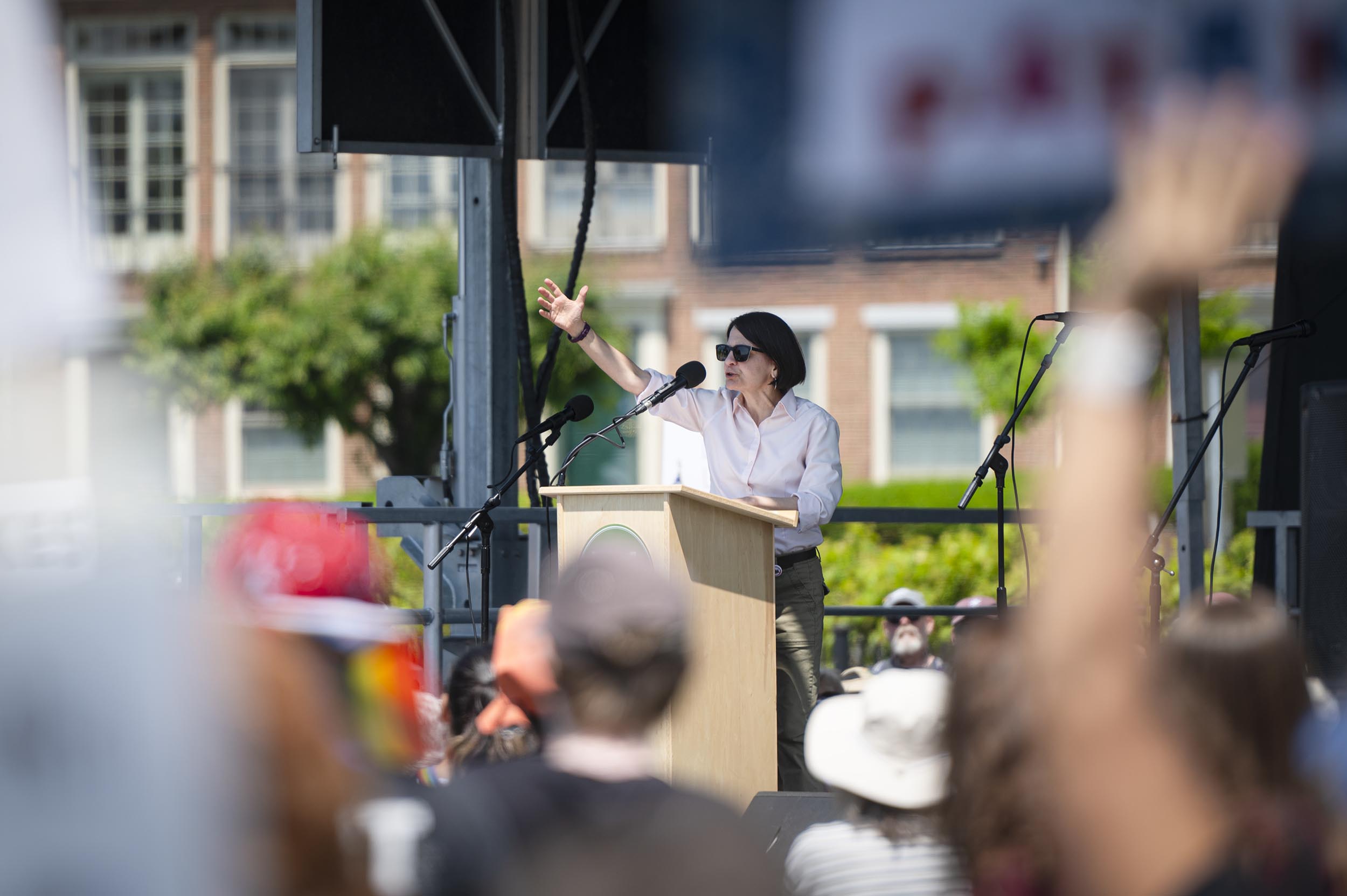 ‘No Kings Day’ draws more than 16,000 to Burlington to protest Trump and authoritarianism A person wearing sunglasses and a hat speaks at a podium on an outdoor stage to a crowd during a daytime event.