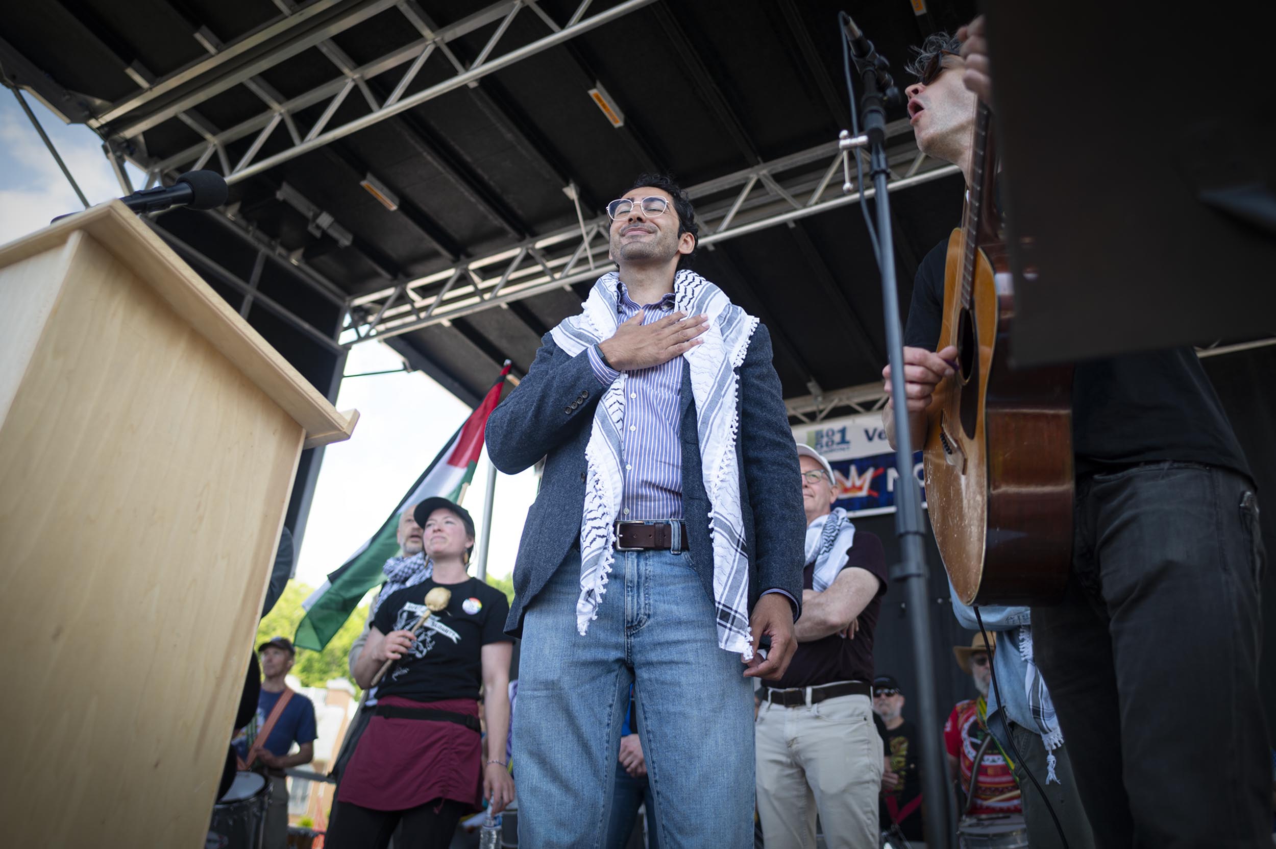 ‘No Kings Day’ draws more than 16,000 to Burlington to protest Trump and authoritarianism A man with a keffiyeh stands on an outdoor stage with his hand on his chest, surrounded by others; a guitar player and podium are also visible.