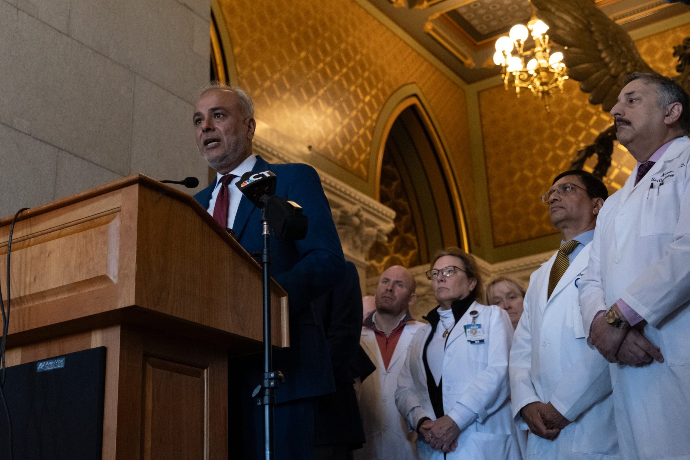 Fix or nix? CT’s ‘certificate of need’ law under scrutiny A man in a suit stands at a podium speaking into a microphone. Doctors in white coats stand behind him.