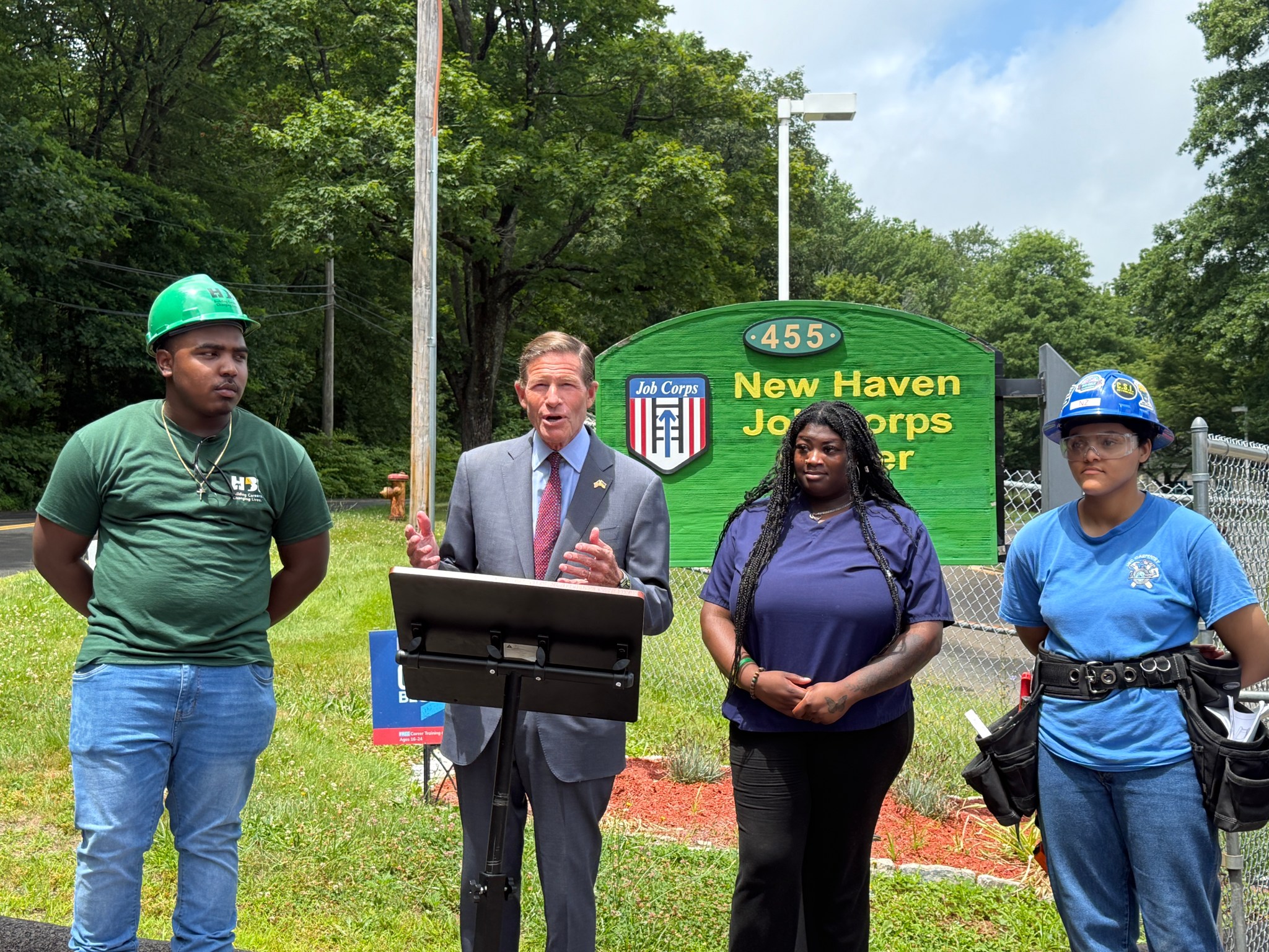 New Haven and Hartford Job Corps centers hang in the balance Senator Blumenthal is standing at a podium in front of the New Haven Job Corps Center sign. Program graduates and enrollees stand to the Senator's left and right side, wearing their vocational gear and uniforms as he speaks in support of Job Corps.