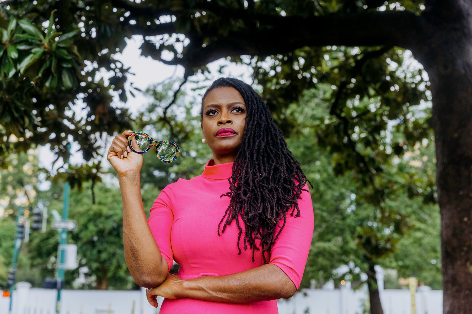 ‘Voting feels like a battle’: A group of Black women reimagines voter turnout Cassandra Welchlin holding glasses in one hand, standing under a tree with a determined expression. She is wearing a bright pink dress, with her long locs draped over her shoulder, and the background features a park setting.