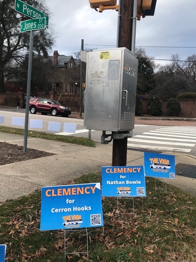 Three blue signs at an intersection, two reading “Clemency for Cerron Hooks” and “Clemeny for Nathan Bowie.” The third reads “The Vigil for Freedom and Racial Justice.”