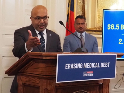 Dev Sangvai gestures with an outstretched palm at a podium labeled "Erasing Medical Debt."