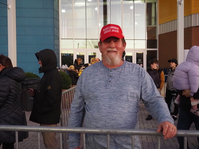 A man in a Make America Great Again hat stands in front of a building in a line with other people.