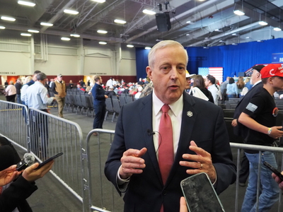 A man in a suit wearing a microphone speaks to a group of reporters in an event center.