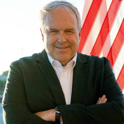 A headshot of a man in a suit standing in front of an American flag.