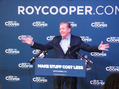 Roy Cooper stands at a podium that reads "Make Stuff Cost Less" with outstretched arms.