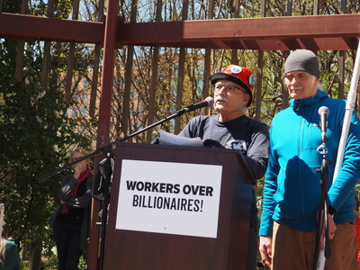 A man in a helmet reads remarks standing at a podium labeled Workers over Billionaires.