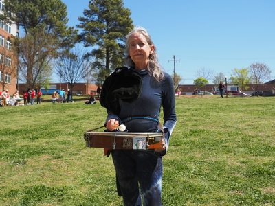 A woman stands holding a drum labeled "No Kings" in a grassy park.