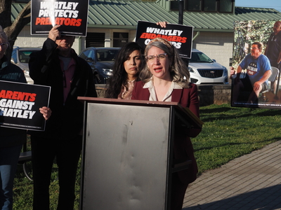 A state senator speaks at a podium with demonstrators behind her holding signs that say "Whatley Protects Predators" and "Parents Against Whatley"