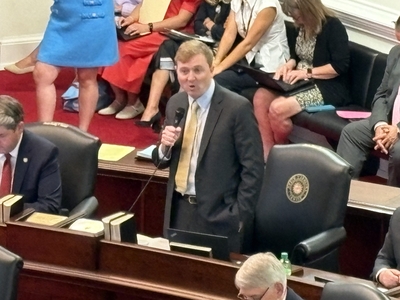 A man speaks into a microphone standing from his chair.