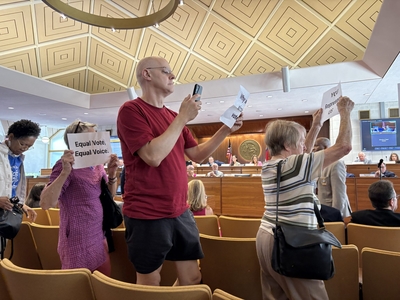 Several protesters walk out of the room holding signs such as "Equal Vote, Equal Voice."