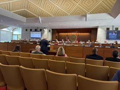 A hearing proceeds before rows of empty chairs.