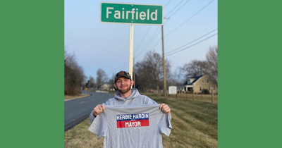 A photo of Herbie Hardin standing in front of a sign for Fairfield, Kentucky. He's holding a t-shirt that says: "Herbie Hardin for Mayor".