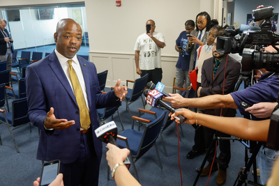 A man in a blue suit and a yellow suit speaks into a stand of microphones.