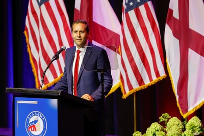 John Wahl, the chair of the Alabama Republican Party, speaks from behind a lectern, wearing a dark blue suit and red tie. A teleprompter is in front of him; the flags of the United States and Alabama stand behind him.