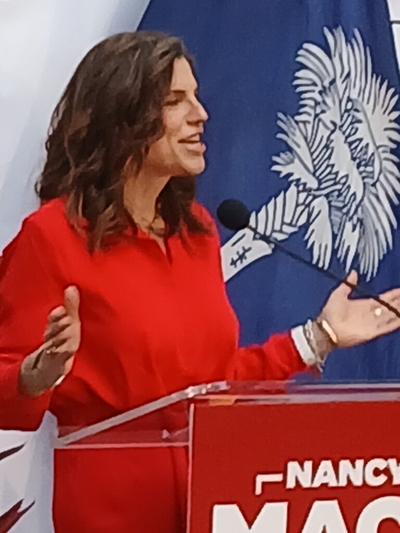 A woman in a red dress at a lectern speaking with her arms etended