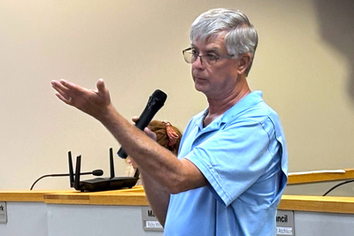 A man in a light blue shirt holding a microphone in his right hand and gesturing with his left arm