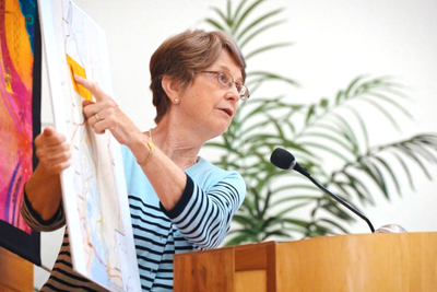 A woman in glasses at a lectern pointing to a chart