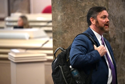 A man in a blue jacket holding a bag and leaning against a pillar
