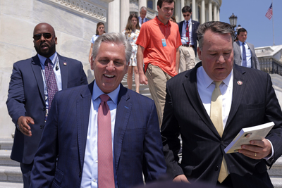 Two men in suits walk down the stairs, one smiling and the other looking down at a pad of paper.