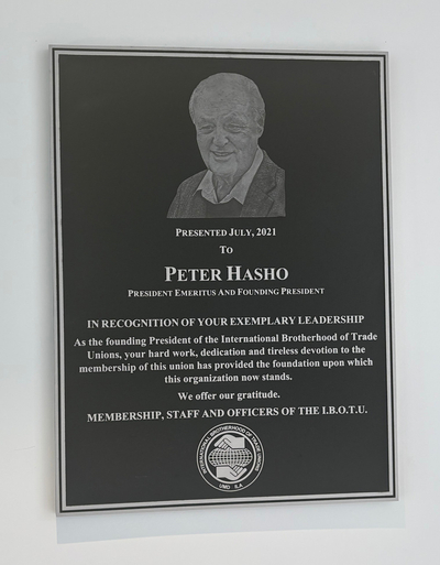 A black and gray plaque mounted on a wall shows Peter Hasho's likeness, stating "Presented July, 2021 to Peter Hasho." The IBOTU logo is on the bottom of the plaque.