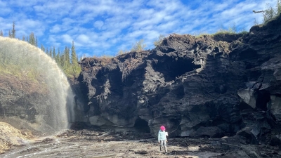 A person walks in a red hat in front of a cliff and a large jet of water, with blue sky behind