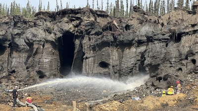 People work in reflective yellow/orange jackets at the base of a cliff, with a high-pressure jet of water spraying across