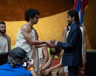 A man in a light-colored tunic, left, receives a rock from a younger man dressed in a blue blazer during a ceremony.