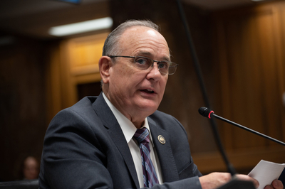 Rep. Bacala sits at a table with a microphone. He is wearing a dark grey suit.