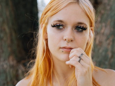 A photo shows a White woman with reddish blonde hair standing in front of some trees. Her left hand is resting against her face.