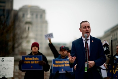 A man in a suit stands in front of a microphone outside.
