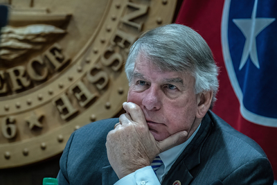 A with gray hair sits before a state seal, his chin propped on his hand.