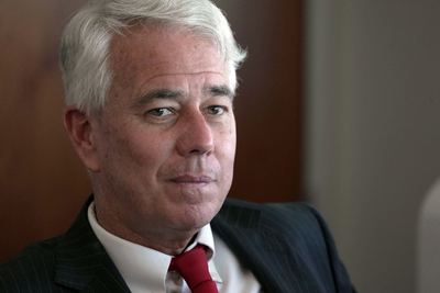 A man in a navy blue suit and red tie, with white hair, smiles slightly.