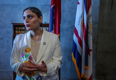 A woman in a white suit with dark hair stands in front of an American flag.
