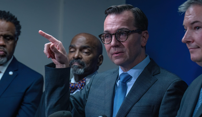 A man in a suit and glasses points his finger while talking.