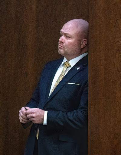 A bald man wearing a dark suit looks thoughtful and clasps his hands.
